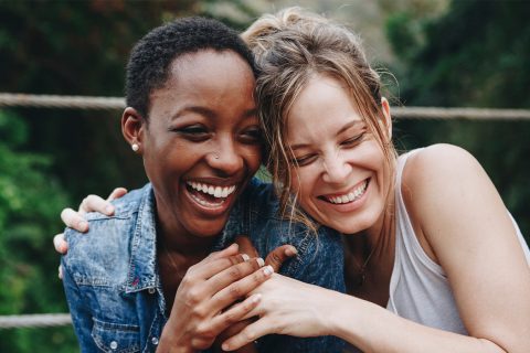 Two women hugging eachother in a close up shot. On the left a black woman wearing a denim jacket and on the right, a caucasion woman wearing a white tank top.