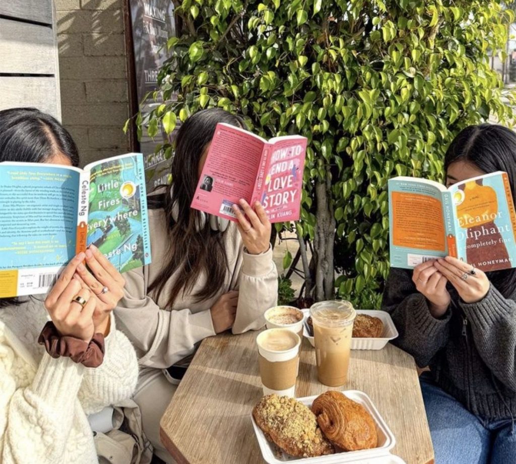 Three friends sit outside of a coffee shop with their book club reads held up in front of their faces.