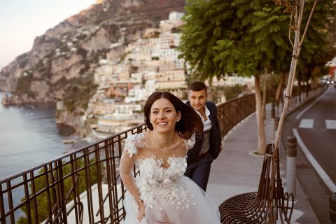 A smiling bride and groom are running down a street in a European location.