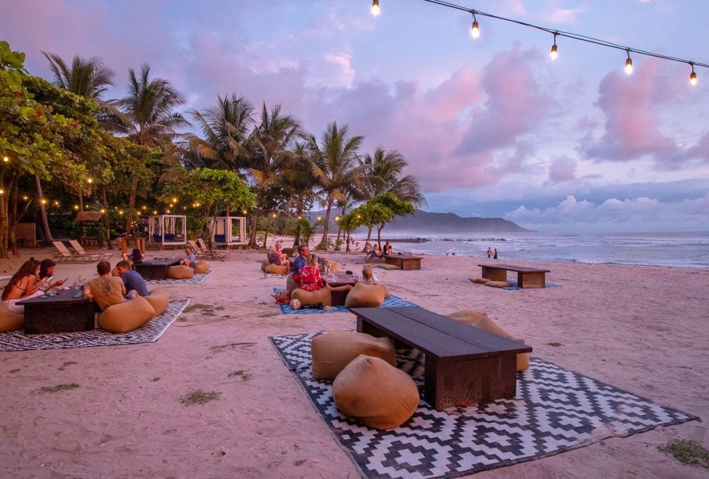 The beachfront area of the Nantipa Hotel in Santa Teresa, Costa Rica, specifically showcasing the open-air seating for Manzú Restaurant with low wooden tables, bean bag seating, and patterned area rugs placed directly on the sand for casual dining.