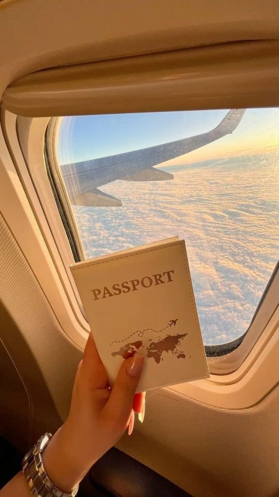 A passport cover decorated with a small world map is held up against an airplane window, showcasing a view of white clouds at sunrise.